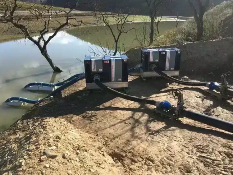 Twee pompsets aan de oever van een waterbassin, met zuigslangen op drijvers in het water en persslangen naar een leiding